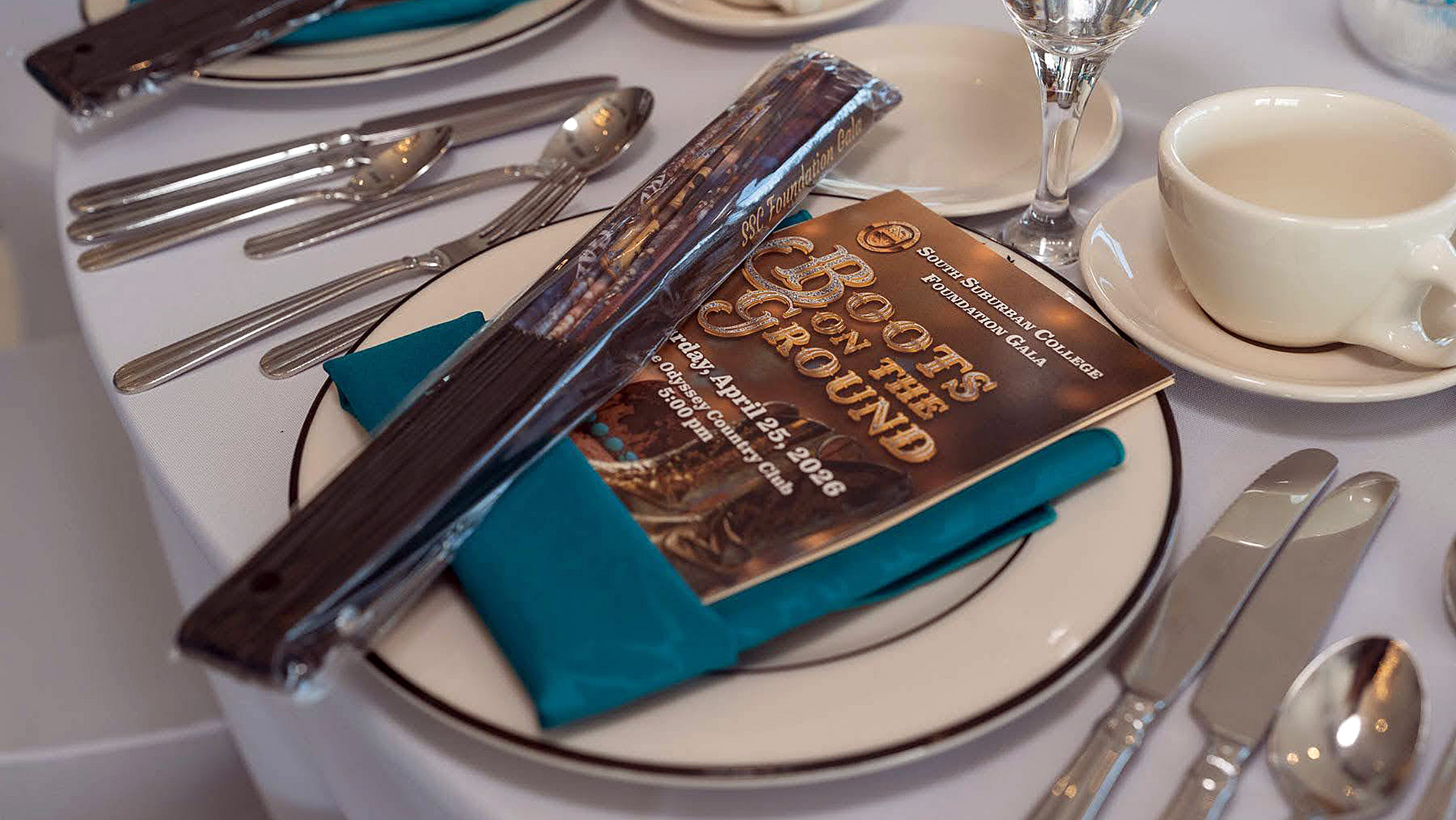 A place setting at the 2026 South Suburban College Foundation Gala, featuring a printed program titled "Boots on the Ground" resting on a white plate with a teal napkin, accompanied by a wrapped party favor and formal silverware.